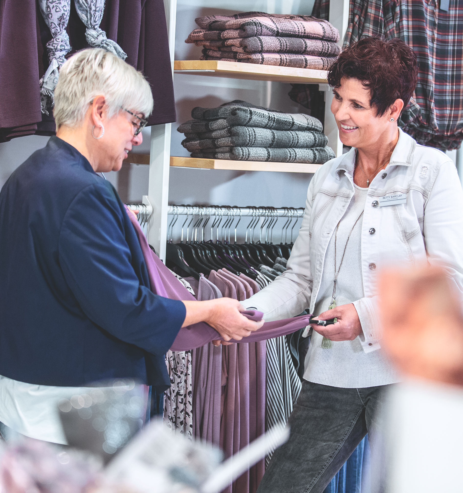 Zwei Frauen stehen in einem Bekleidungsgeschäft. Die eine hält ein lila Kleidungsstück hoch, während die andere es begutachtet. Im Hintergrund sind Regale mit gefalteter Kleidung zu sehen. Beide scheinen in ein freundliches Gespräch vertieft zu sein.