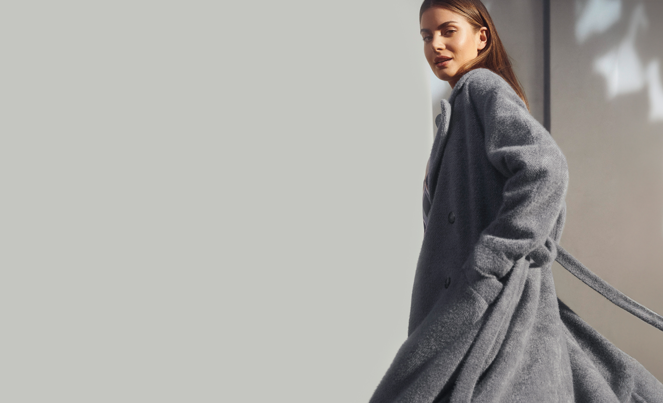 Eine Frau trägt einen langen, flauschigen Mantel in Grau und blickt über die Schulter. Der elegante Look wird durch die fließende Bewegung des Mantels unterstrichen.